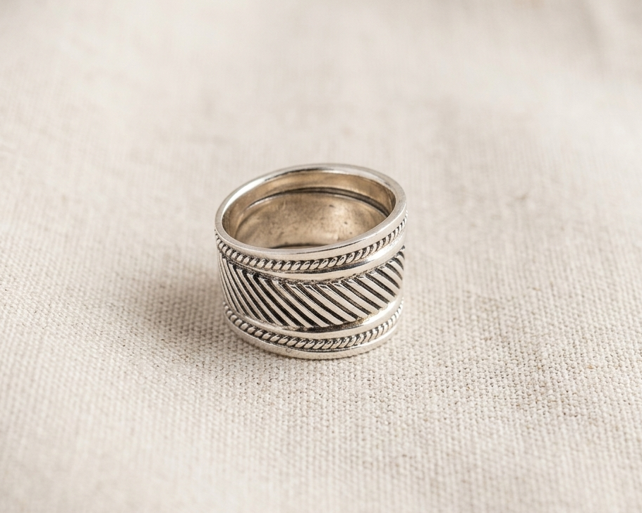 Substantial sterling silver wide band ring featuring an intricate oxidized diagonal woven pattern bordered by delicate rope-twist edges, displayed on a textured light linen background.