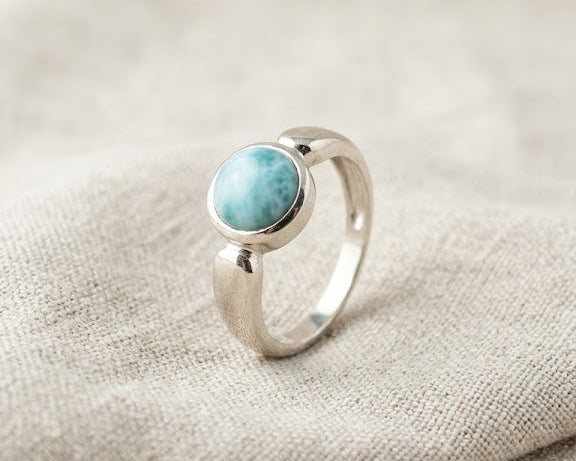 Sterling silver bezel ring featuring a round cabochon-cut natural blue Larimar gemstone on a light beige linen fabric background.