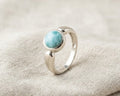 Sterling silver bezel ring featuring a round cabochon-cut natural blue Larimar gemstone on a light beige linen fabric background.
