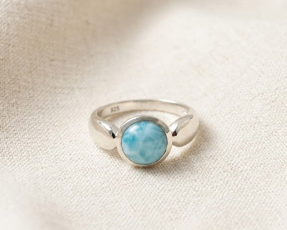 Sterling silver bezel ring featuring a round cabochon-cut natural blue Larimar gemstone on a light beige linen fabric background.
