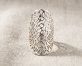 A handcrafted 925 sterling silver statement ring featuring an intricate organic filigree cutout design, shown standing in a top-down view on a light oatmeal-colored linen background under professional studio lighting