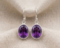 Close-up photograph of a pair of genuine oval-cut purple amethyst drop earrings in polished sterling silver bezel settings.