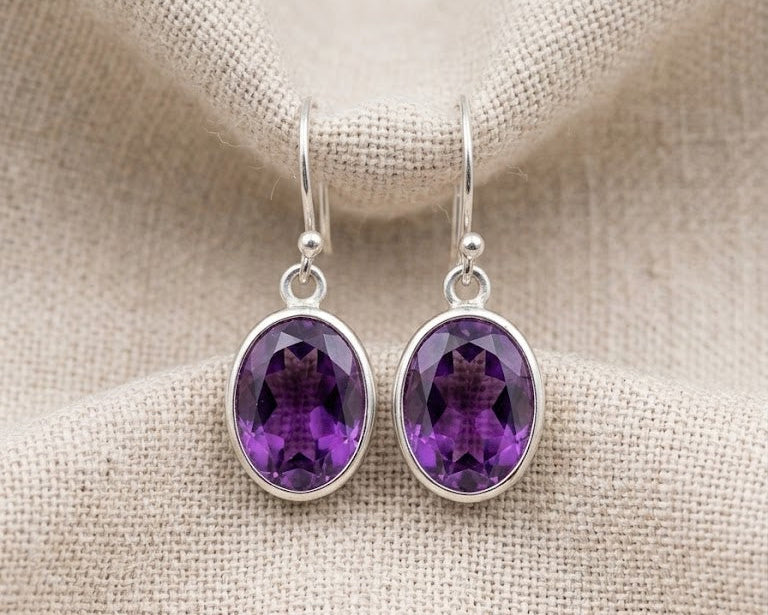 Close-up photograph of a pair of genuine oval-cut purple amethyst drop earrings in polished sterling silver bezel settings.