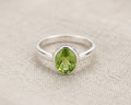 A close-up photograph captures a single sterling silver ring featuring a vibrant, faceted green peridot stone in an oval setting. The ring is centered on a textured, light beige linen fabric background, showcasing the jewelry's simple and elegant design.