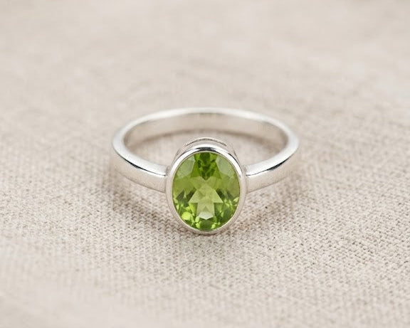 A close-up photograph captures a single sterling silver ring featuring a vibrant, faceted green peridot stone in an oval setting. The ring is centered on a textured, light beige linen fabric background, showcasing the jewelry's simple and elegant design.