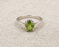 Detailed close-up image of a sterling silver peridot solitaire ring with a vibrant green, oval-cut peridot in a four-prong setting, resting on a light beige textured linen fabric background.