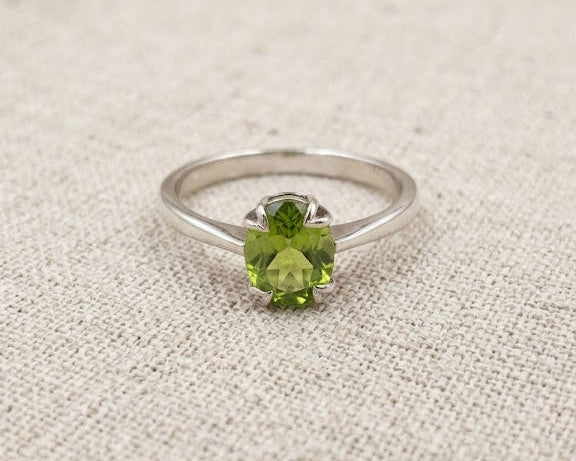 Detailed close-up image of a sterling silver peridot solitaire ring with a vibrant green, oval-cut peridot in a four-prong setting, resting on a light beige textured linen fabric background.